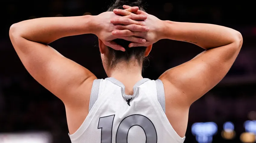 Kelsey Plum holds her hands behind her head, interlocking her fingers. She is wearing a white and silver jersey with the number 10 on it and the name "Plum" underneath it.