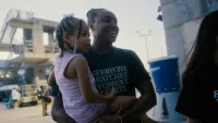 Olympian Simone Manuel wears a shirt that says "Everyone watches women's sports" and holds a young Black girl in her arms as they enter a pool area
